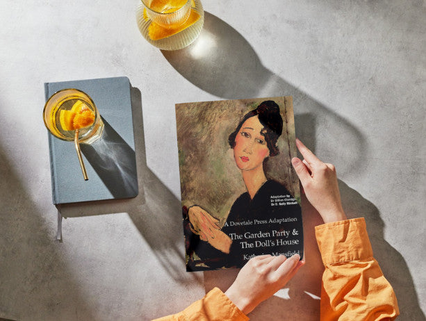 An overhead shot of a woman arms on a grey marble table. She's holding a copy of The Garden Party and The Doll's House. There's a jug and glass of orange water on a grey notebook beside her.