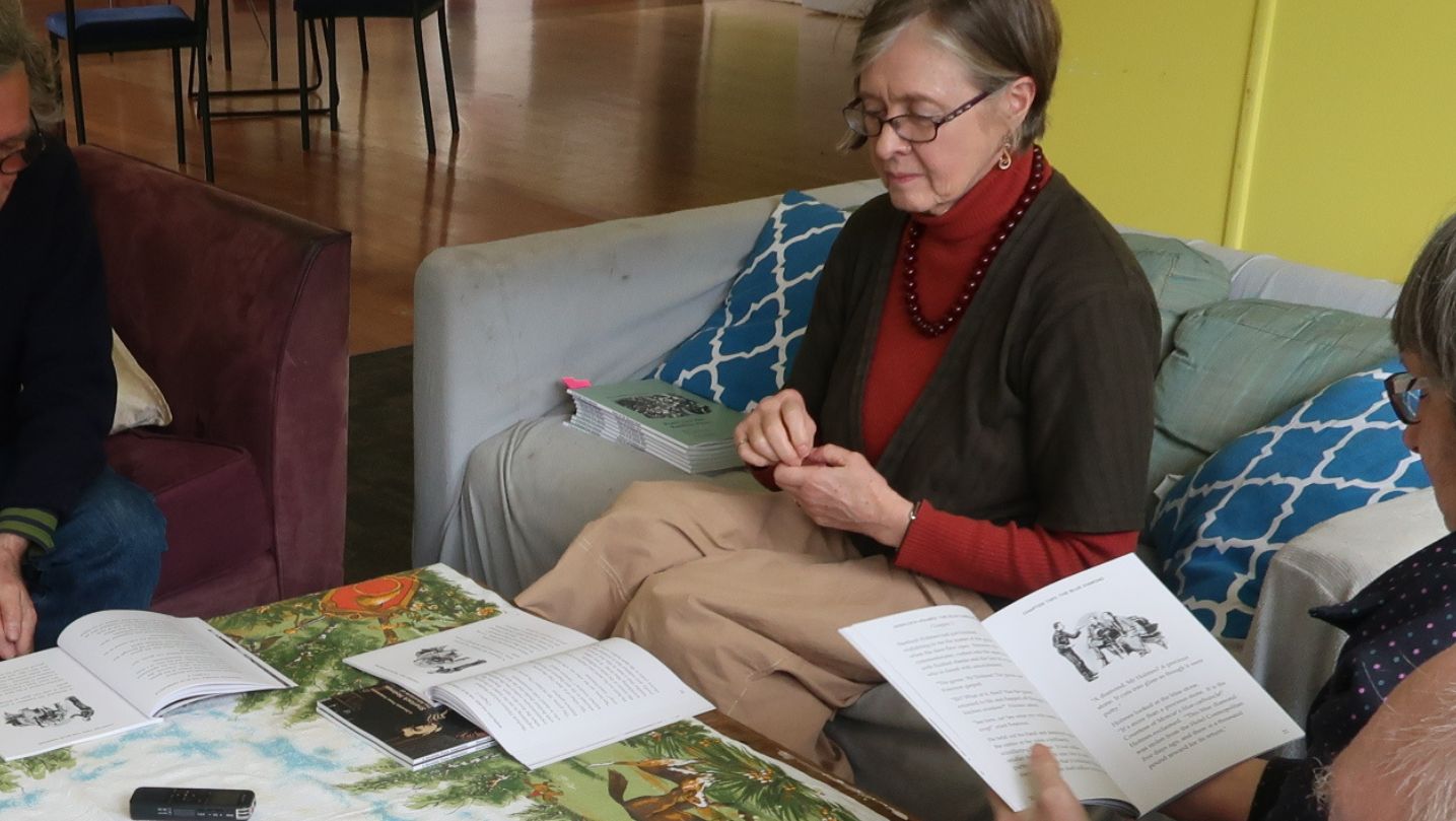 Gill leading a book group in a community centre. Two books are open on the table and a person holds one book open
