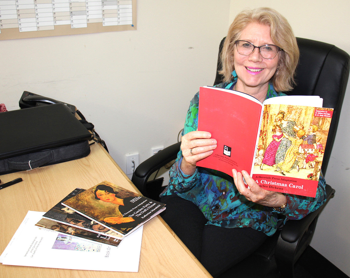 Dr Sally Rimkeit sitting and reading A Christmas Carol at her desk. She smiles warmly at the camera.